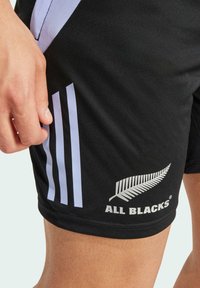 Musta urheilu-shortsi, joissa on laventelinvärisiä raidoja, valkoisella saniainenlogolla ja "ALL BLACKS" -tekstillä. Valmistettu sileästä, venyvästä kankaasta.