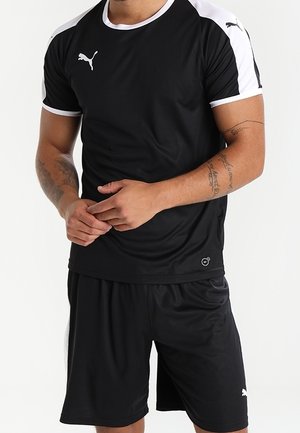 Mann trägt schwarze und weiße Puma-Sportbekleidung, bestehend aus einem kurzärmeligen Shirt und Shorts, und steht mit vor dem Körper verschränkten Händen.