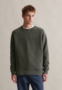 Zielona bluzka z bawełny, z okrągłym dekoltem i rękawami raglanowymi. Subtelne logo z przodu, w połączeniu z niebieskimi dżinsami.