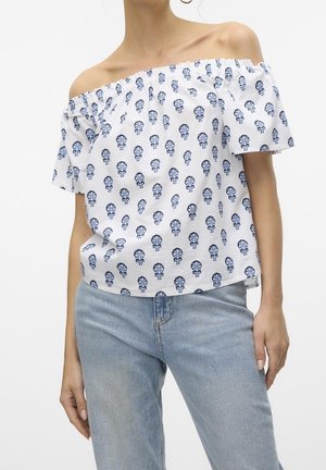 Vrouw met witte off-shoulder blouse met blauw bloemenprint en lichtblauwe spijkerbroek, staand tegen een effen witte achtergrond.