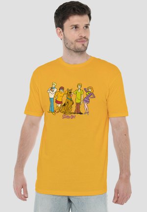Man wearing a yellow T-shirt with Scooby-Doo and four characters standing around the dog in colorful outfits.