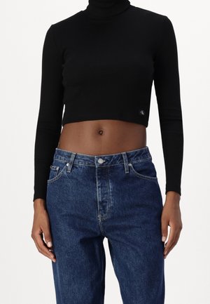Femme portant un top court noir à col roulé manches longues et un jean bleu taille haute, debout les mains détendues sur les côtés.