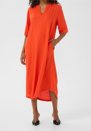 Kvinde iført en løs, lys orange midi-kjole med albuelange ærmer og sidelommer, kombineret med sorte spidse hæle.