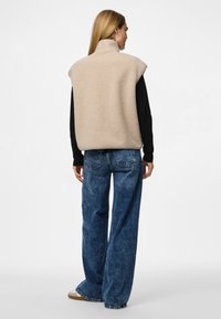 Vestă bej din fleece cu guler înalt și design fără mâneci, purtată peste un top negru cu mânecă lungă, asortată cu blugi din denim albastru cu croială largă.