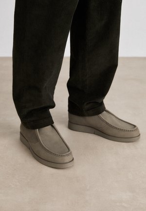 Persona con pantalones de pana negros y mocasines de piel beige sin cordones, de pie sobre un suelo claro.