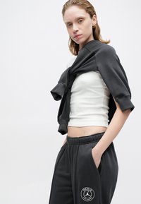 Biały bawełniany top na ramiączkach, noszony pod ciemnoszarym swetrem zarzuconym na ramiona, w połączeniu z szarymi dresami z białym logo na udzie.