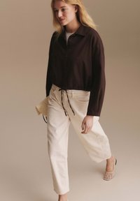 Blonde Frau, die beige Hosen mit weitem Bein, eine schwarze Jacke mit Knöpfen und geflochtene Mules trägt, während sie eine kleine beige Handtasche hält.