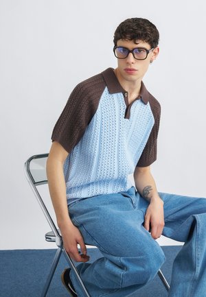 Fiatal férfi göndör sötét hajjal és szemüveggel, barna és világoskék kötött galléros pólót, illetve bő kék farmert viselve, fém széken ül.