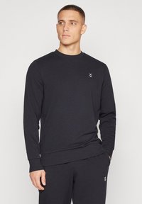 Homme portant un sweat-shirt noir à manches longues et un pantalon assorti, debout avec une main dans la poche, regardant vers la gauche sur fond blanc.