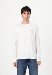 Witte longsleeve t-shirt met een klein zwart logo op de linkerkant van de borst, gemaakt van zachte stof. Draagt met blauwe denim jeans.