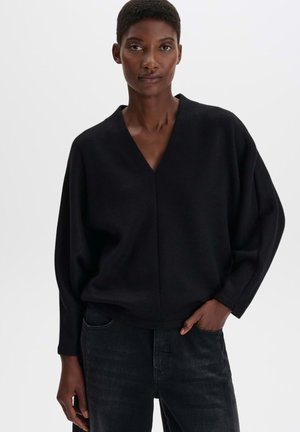 Woman with short hair wearing a black V-neck sweater and dark jeans, standing with one hand in pocket against a plain background.