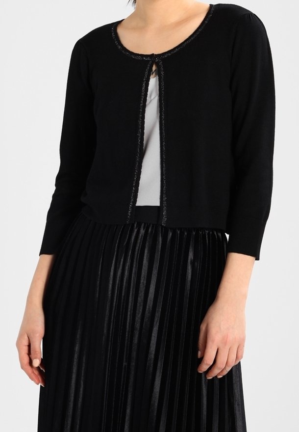 Femme portant un cardigan court noir orné de paillettes par-dessus un haut blanc et une jupe plissée noire taille haute.