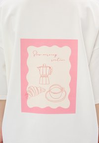 Witte T-shirt met een roze geschulpte graphic met lijntekeningen van een koffiepot, croissant en mok. Tekst luidt "Trage ochtendroutine."