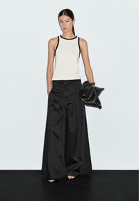 White tank top with black trim, paired with wide-leg black pants. Holding a textured black bag, stance neutral, against a light background.