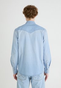 Camisa de mezclilla azul claro con mangas largas, cuello puntiagudo y cierres de botones a presión. Presenta un diseño de yugo y costuras decorativas en la parte posterior.