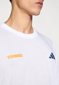 Camiseta blanca de algodón con cuello redondo, presenta texto naranja "TENNIS" en el pecho izquierdo y un logo de Adidas azul marino en el hombro derecho.