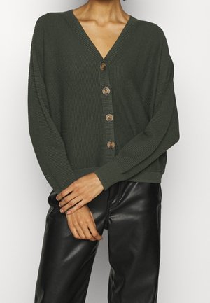 Cardigan en maille vert foncé avec quatre boutons en bois porté avec un pantalon en cuir noir, les mains de la personne croisées à la taille.