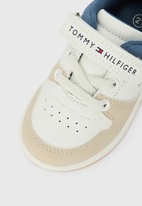 Zapatilla blanca y beige con parte superior de cuero texturizado, puntera redonda, sistema de cordones y una correa de velcro con la etiqueta "TOMMY HILFIGER".