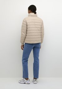 Beige gewatteerde jas met een horizontaal gestikt patroon, hoge kraag en ribgebreide manchetten. Gecombineerd met blauwe jeans en witte sneakers.