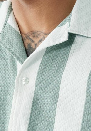 Camicia a righe verdi e bianche con una trama testurizzata, colletto corto e chiusura con un solo bottone al collo, che rivela un tatuaggio sottostante.