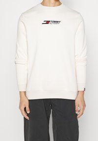 Pullover color crema con texture a coste, con logo in rosso, bianco e nero. Maniche lunghe e scollo a rotondo.
