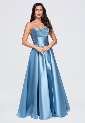 Femme portant une robe longue, bleu clair et brillante, sans bretelles, avec un col orné de perles, debout, les mains jointes devant un fond clair uni.