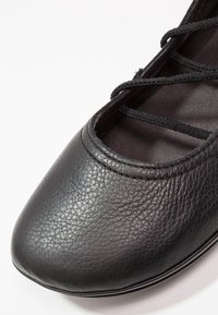 Gros plan d'une ballerine en cuir noir texturé avec des brides élastiques croisées et un bout arrondi.