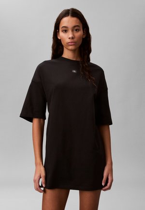 Jeune femme aux longs cheveux foncés portant une robe t-shirt noire oversize Calvin Klein sur un fond gris uni.