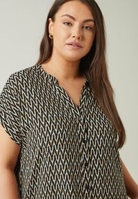 Blusa sin mangas en negro y crema con un patrón de zigzag, que incluye un cuello y una placket de botones. Tela suave con un ajuste suelto y relajado.
