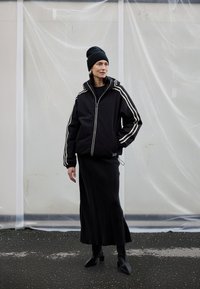 Črna puhovka s belimi črtami, črna dolga krila in črna pletena kapa. Model nosi črne škornje z peto, minimalističen dizajn.