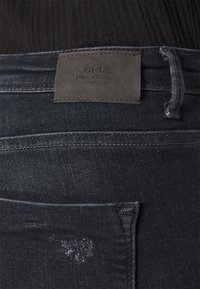 Vaqueros de mezclilla oscura con un tejido texturizado. Presentan un parche de cuero con el logo en la cintura y sutiles detalles deslavados. Un solo bolsillo trasero visible.