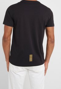 Black cotton t-shirt featuring a round neckline and short sleeves, with a gold striped accent near the hem on the back.