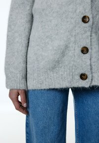 Grijze wollen cardigan met een zachte textuur, voorzien van grote bruine knopen en ribbelmanchetten. In combinatie met blauwe spijkerbroek.