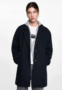 Marinrandig kappa med skräddarsydd passform, breda slag och två knappar. Lagd över en grå zip-up hoodie under, som visas med fickor.