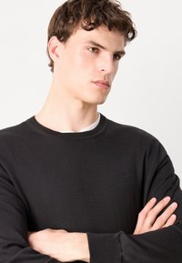 Pull noir à manches longues avec un col et des poignets côtelés, présentant un petit logo sur la poitrine, fabriqué en tissu mélangé coton.
