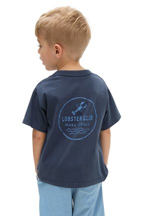 Blonder Junge im Profil, der ein dunkelblaues "Lobster Club" T-Shirt und hellblaue Shorts vor weißem Hintergrund trägt.