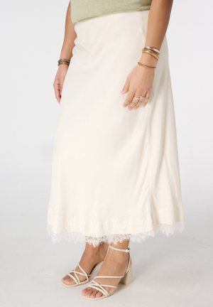 Vrouw draagt een crèmekleurige midi-rok met kanten zoom, gecombineerd met beige sandalen met blokhakken en bandjes, en gouden armbanden aan beide polsen.