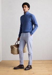 Μπλε πουλόβερ πλεκτό με υψηλό γιακά, ανοιχτό γκρι παντελόνι, καφέ μοκασίνια και μπεζ τσάντα με καφέ λεπτομέρειες. Μινιμαλιστικό φόντο.