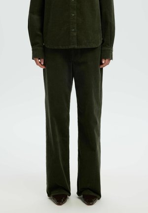 Personne portant un pantalon large en velours côtelé vert foncé et une chemise assortie boutonnée, debout avec les bras détendus le long du corps.