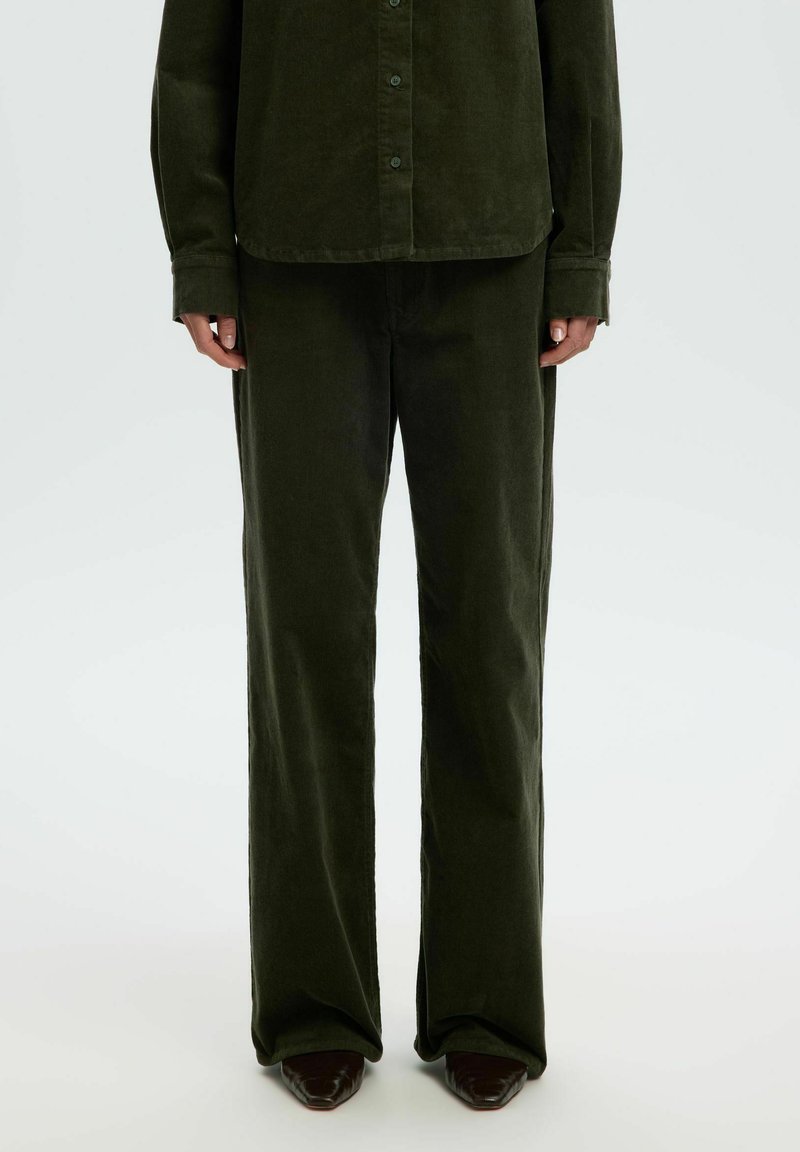 Personne portant un pantalon large en velours côtelé vert foncé et une chemise assortie boutonnée, debout avec les bras détendus le long du corps.