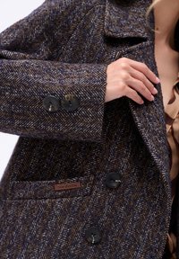 Ręka dostosowująca ciemnoniebieską i brązową marynarkę w wzór jodełki z czarnymi guzikami i metką "blue shadow" na kieszeni.