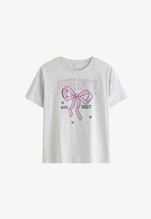 Teenagepige med langt blondt hår iført en grå T-shirt med et pink sløjfe-design, sort nederdel, hvide sokker og sølvfarvede sneakers.