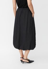 Donna che indossa una gonna midi nera stropicciata con vita elastica, top nero e scarpe slingback nere a tacco basso, in piedi con le mani nelle tasche.