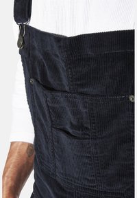 Marineblau Corduroy-Latzhose mit verstellbaren Trägern, ausgestattet mit Vordertaschen und strukturiertem Stoff. Nahaufnahme, die die Nähte und Beschläge hervorhebt.