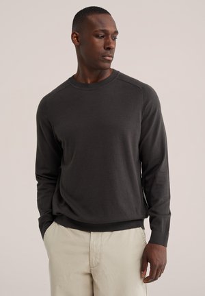 Homme portant un pull à manches longues gris foncé et un pantalon beige clair, debout les mains dans les poches, regardant sur le côté sur un fond uni.