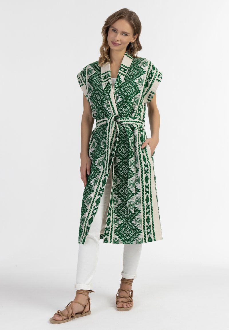 Zaļš un balts rakstots garais veste ar īsām piedurknēm, jostu vidukļa līmenī un kabatām, apvienots ar baltām biksēm un brūnām siksniņu sandalēm.