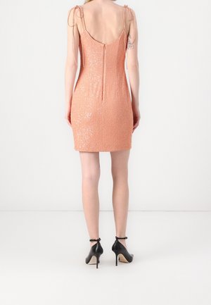 Kobieta w bezrękawnikowej, brzoskwiniowej sukience mini z cekinami i wiązanymi ramiączkami oraz czarnych szpilkach z paskiem wokół kostki, stojąca na białym tle.