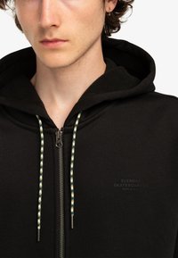 Black zip-up hoodie with a soft texture, featuring a drawstring with green and white pattern, and printed label on the chest.