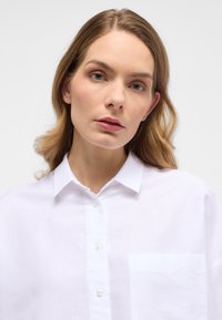 Camicia bianca con bottoni realizzata in tessuto leggero, caratterizzata da un colletto allargato e una tasca sul petto, con una vestibilità rilassata e una texture morbida.