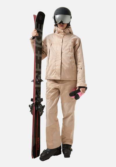 Skikleding in lichtbeige materiaal, bestaande uit een jas met ritssluiting en een broek. Accessoires zijn onder andere zwarte ski's met rode accenten en roze skischoenen.